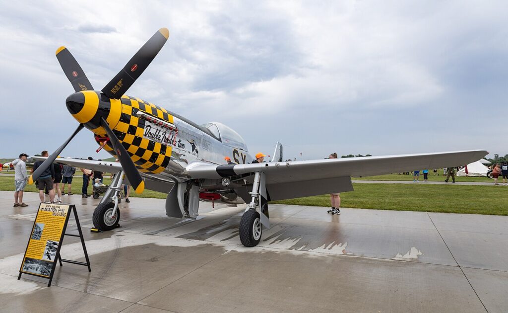 Restored P-51 Mustang fighter aircraft on display at an airshow, featuring a yellow-and-black checkered nose and polished aluminum finish.